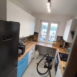 SZAR-BUD Firma remontowo budowlana Sp.z o.o. - Kuchnia po remoncie: białe szafki z drewnianym blatem, niebieskie fronty dolne, wzorzysta ściana nad blatem. Widoczny odkurzacz i narzędzia, co sugeruje końcowy etap prac.