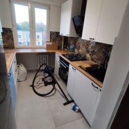 SZAR-BUD Firma remontowo budowlana Sp.z o.o. - Kuchnia po remoncie: białe szafki z czarnymi uchwytami, drewniany blat, wzorzysta ściana nad blatem. Na podłodze odkurzacz i worek. Widok z okna na budynki.