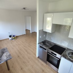 SZAR-BUD Firma remontowo budowlana Sp.z o.o. - Jasne wnętrze po remoncie: białe ściany, panele podłogowe, nowoczesna kuchnia z marmurowym blatem i białymi szafkami. Widoczne ślady po malowaniu w pokoju.