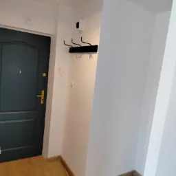 Korytarz po remoncie: drewnopodobne panele podłogowe, pomalowane na biało ściany z zamontowanym czarnym wieszakiem na ubrania, ciemne drzwi wejściowe z złotą klamką oraz sufitowe oświetlenie punktowe.