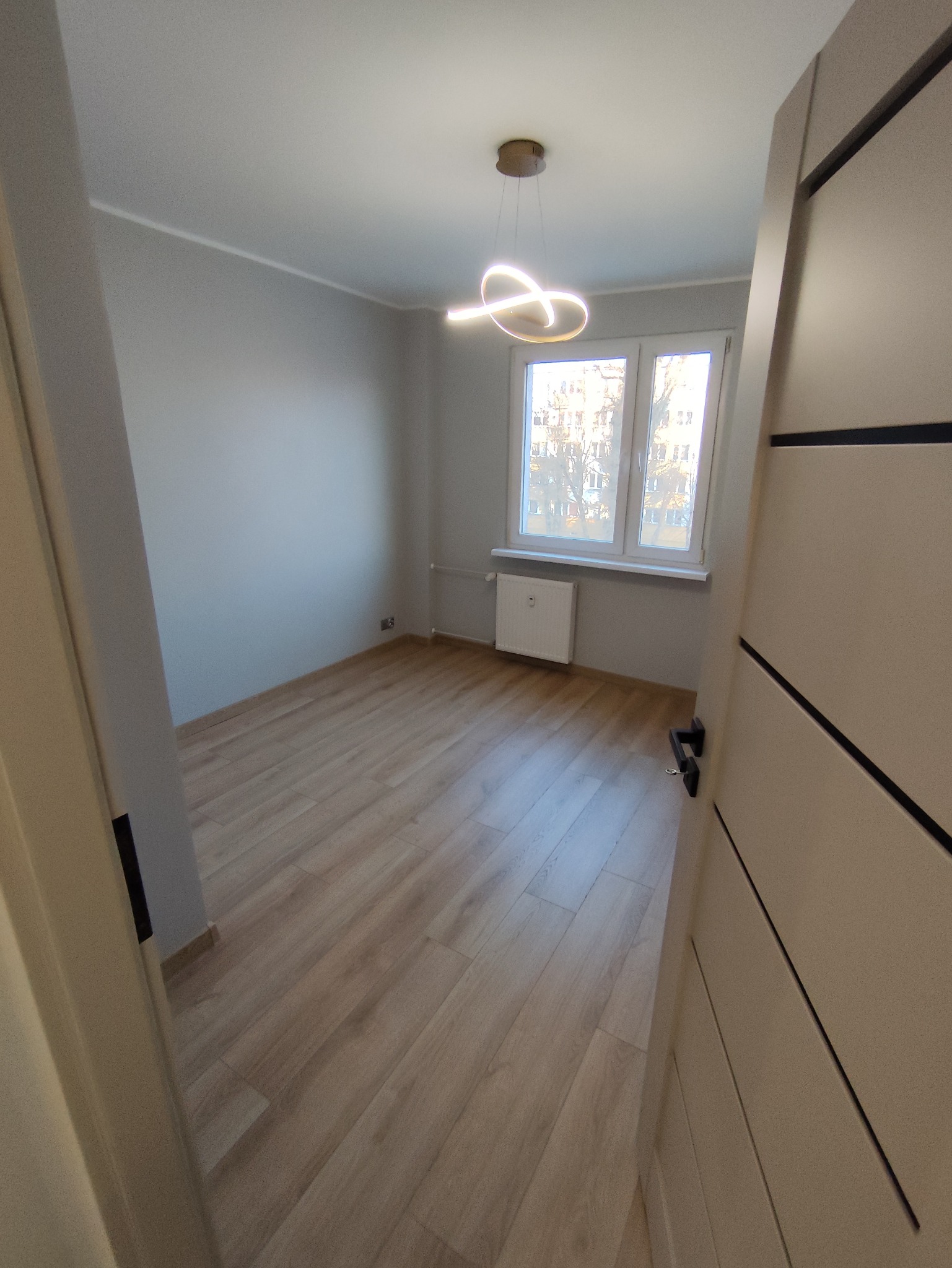 Jasne panele podłogowe w pokoju z szarymi ścianami i nowoczesną lampą. Widok zza uchylonych drzwi, okno z widokiem na budynki w tle.