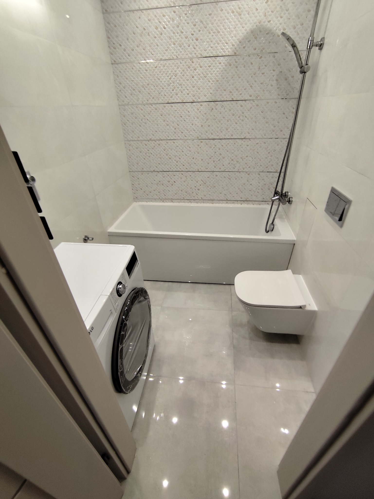 Jasna łazienka po remoncie: białe płytki z delikatnym wzorem, wanna, toaleta podwieszana i pralka. Nowoczesne wykończenie, minimalistyczny styl.