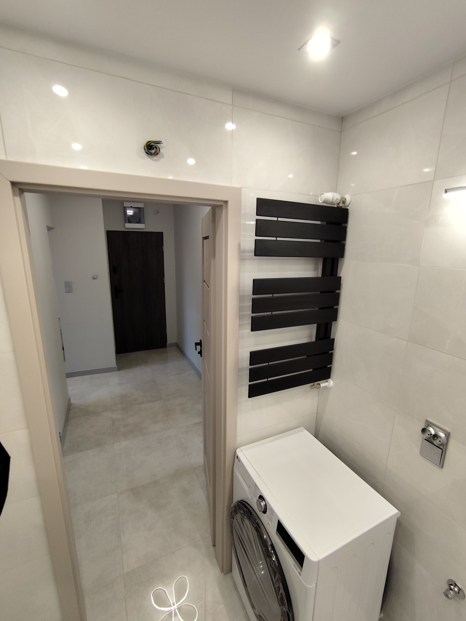 Biała łazienka z pralką LG, czarnym grzejnikiem drabinkowym i błyszczącymi płytkami. Widok na korytarz przez otwarte drzwi. Minimalistyczny design.