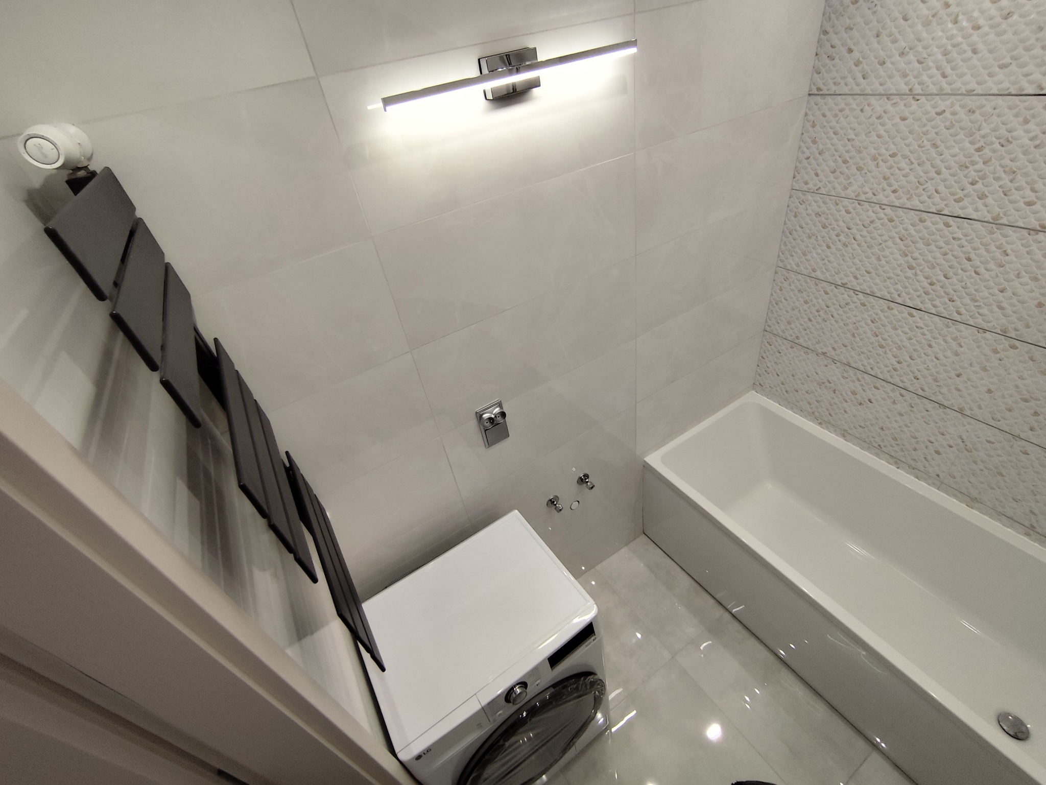Nowoczesna łazienka po remoncie: białe płytki, dekoracyjna ściana, pralka LG, grzejnik drabinkowy i oświetlenie LED. Widok z góry.