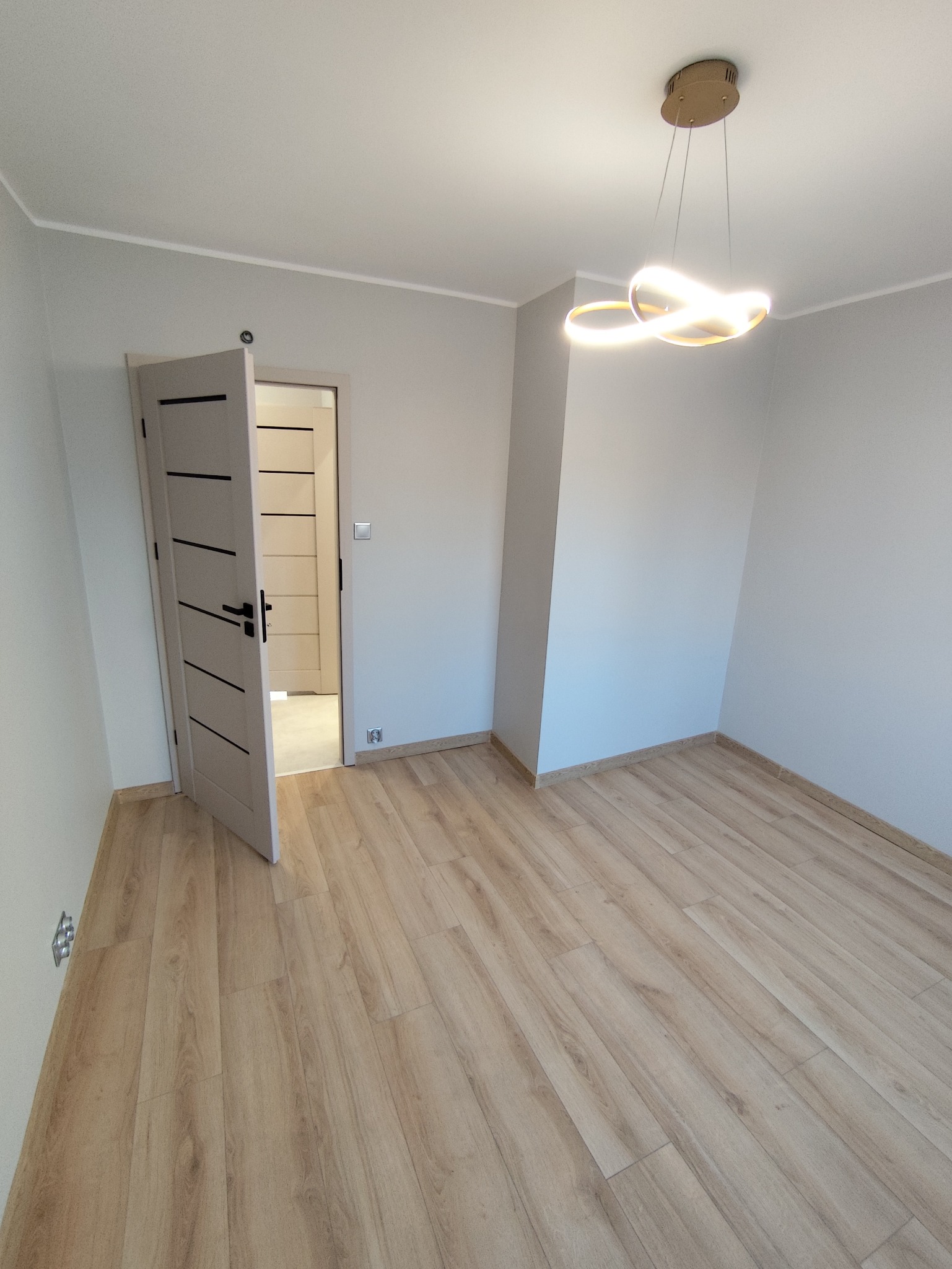 Jasne panele podłogowe w pustym pokoju z otwartymi drzwiami i nowoczesną lampą sufitową. Świeżo wykończone wnętrze w neutralnych barwach.