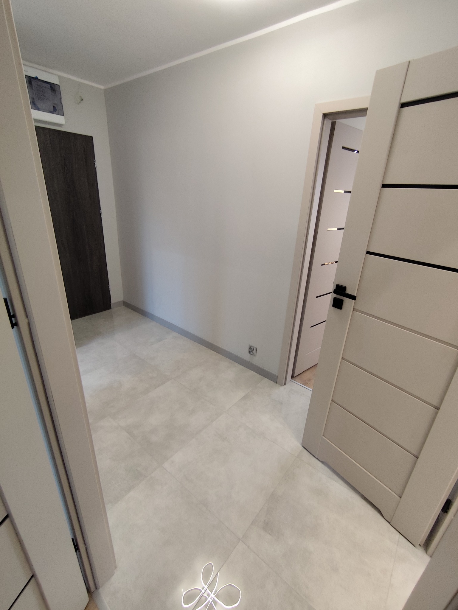 Jasne, świeżo wykończone wnętrze z szarymi płytkami podłogowymi i dwiema parami drzwi w odcieniach beżu. Widoczne listwy przypodłogowe i gładkie, pomalowane ściany.