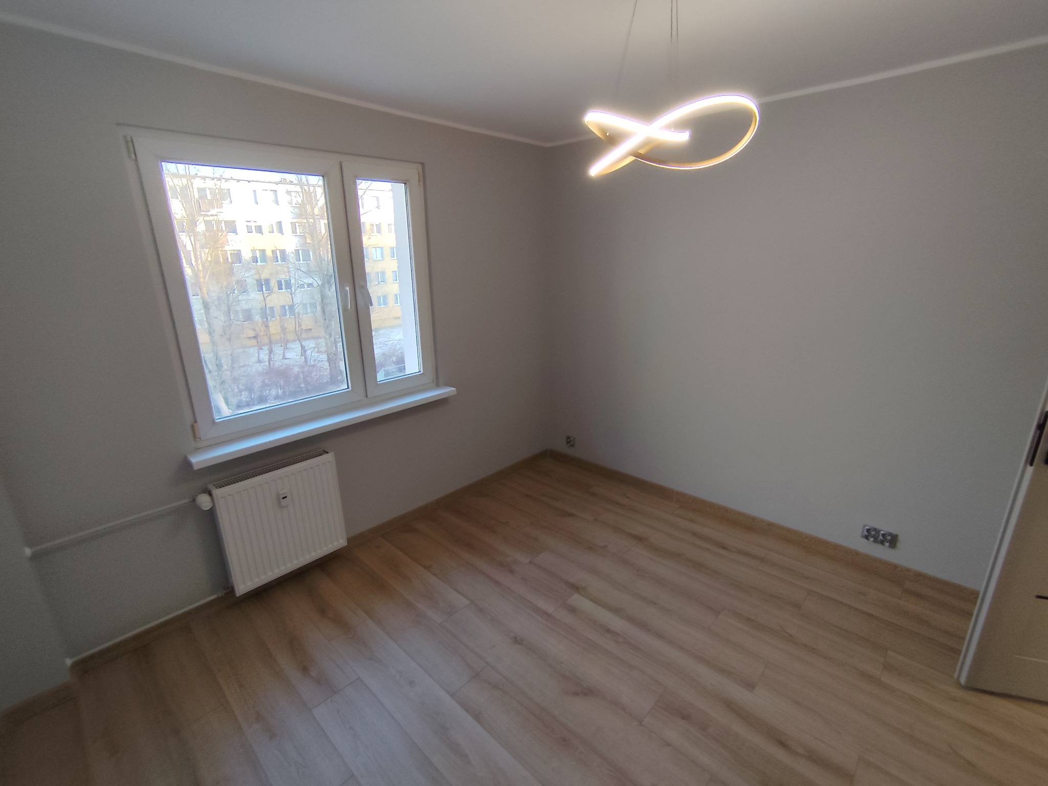 Jasne panele podłogowe w pokoju po remoncie, szare ściany, białe okno z widokiem na blok, nowoczesna lampa sufitowa. Widoczne gniazdka elektryczne i grzejnik.