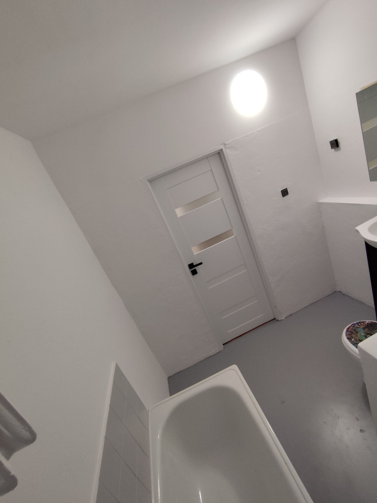 Biała łazienka po remoncie z wanną, umywalką i drzwiami z przeszkleniami. Minimalistyczny design, jasne oświetlenie, szara podłoga. Widok na całe pomieszczenie.