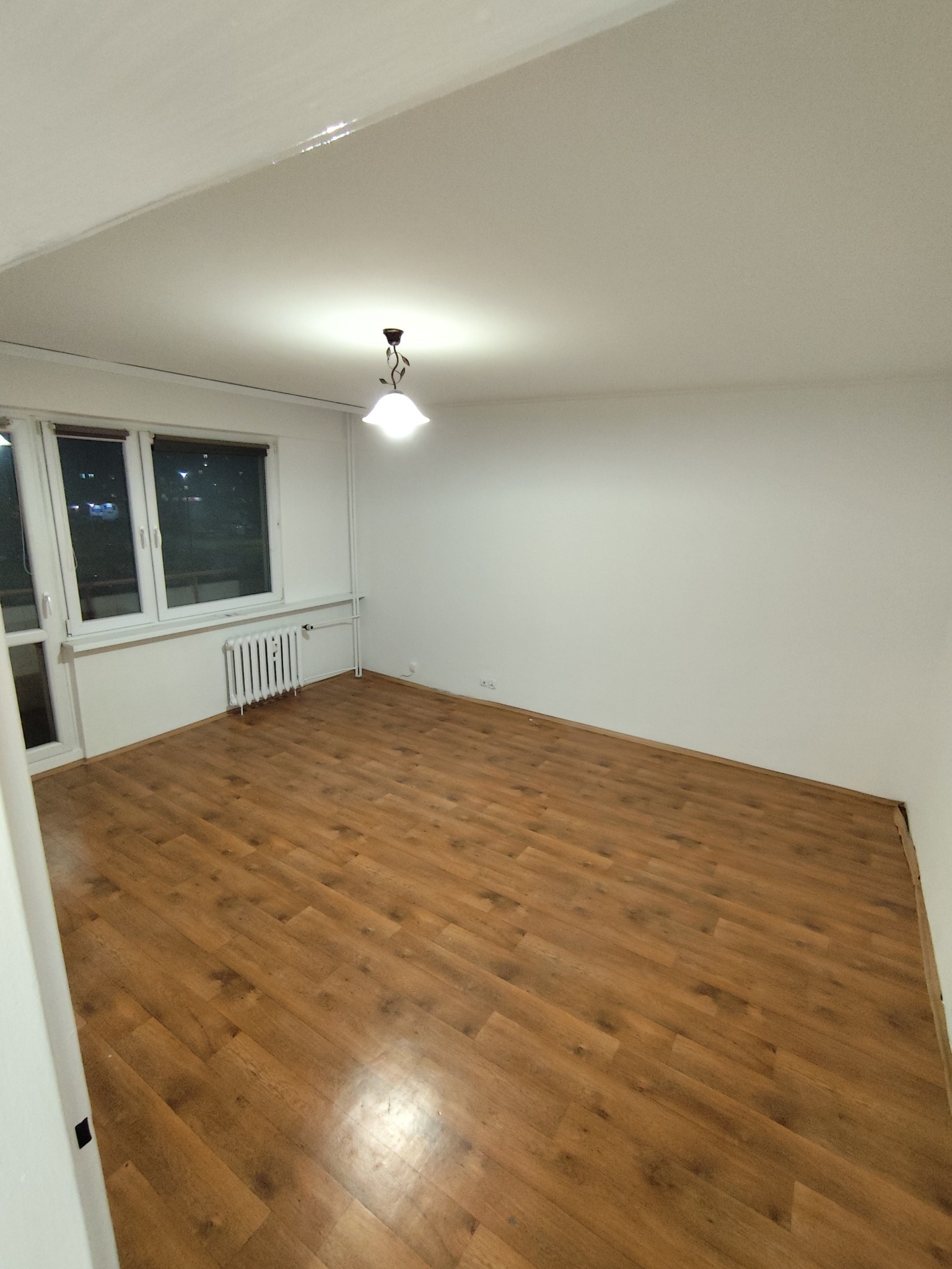 Pokój po remoncie: brązowe panele podłogowe, białe ściany, okno z grzejnikiem. Widok na całe wnętrze z perspektywy wejścia, oświetlone centralną lampą.