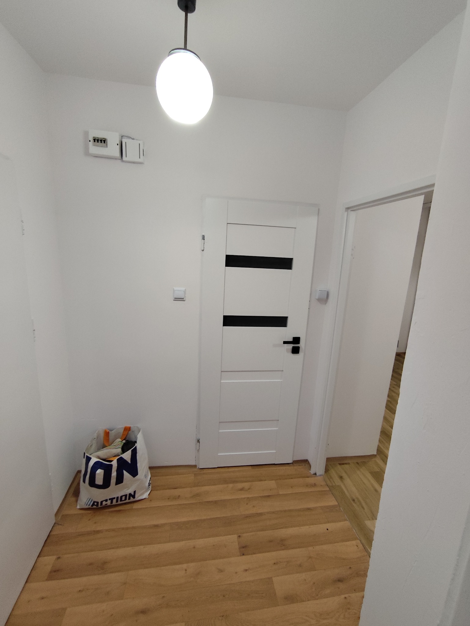Jasne panele podłogowe w korytarzu po remoncie w Warszawie. Widoczne białe ściany, drzwi z czarnymi wstawkami i torba na podłodze. Minimalistyczny design.