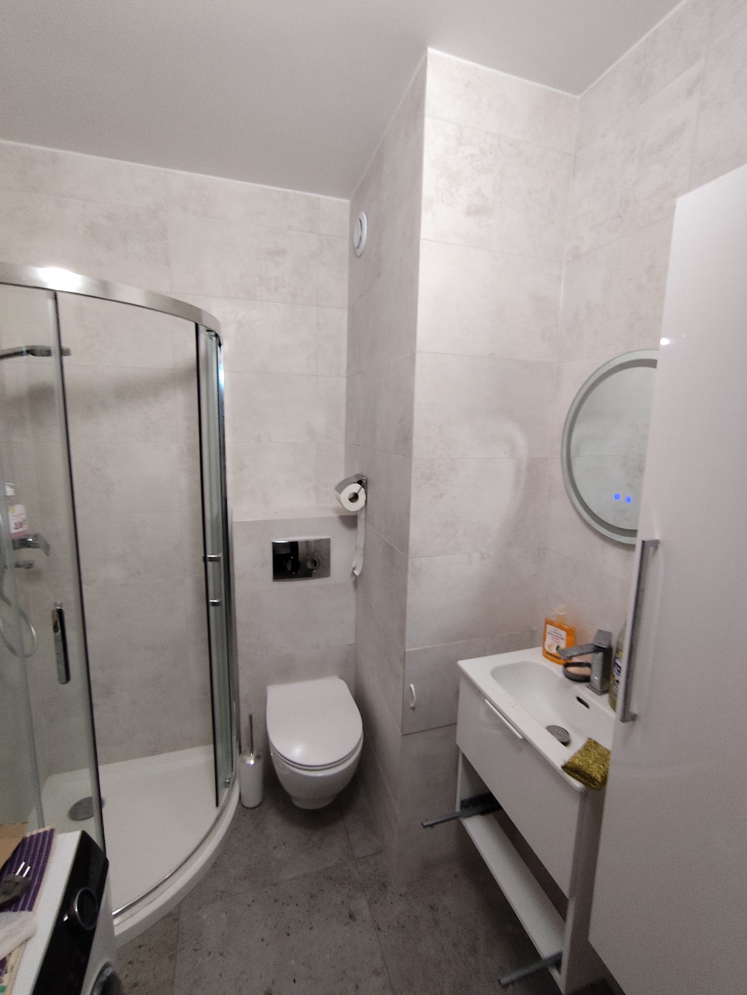 Jasna łazienka po remoncie: kabina prysznicowa z zaokrąglonymi drzwiami, podwieszana toaleta, umywalka z szafką i okrągłe lustro z podświetleniem LED.