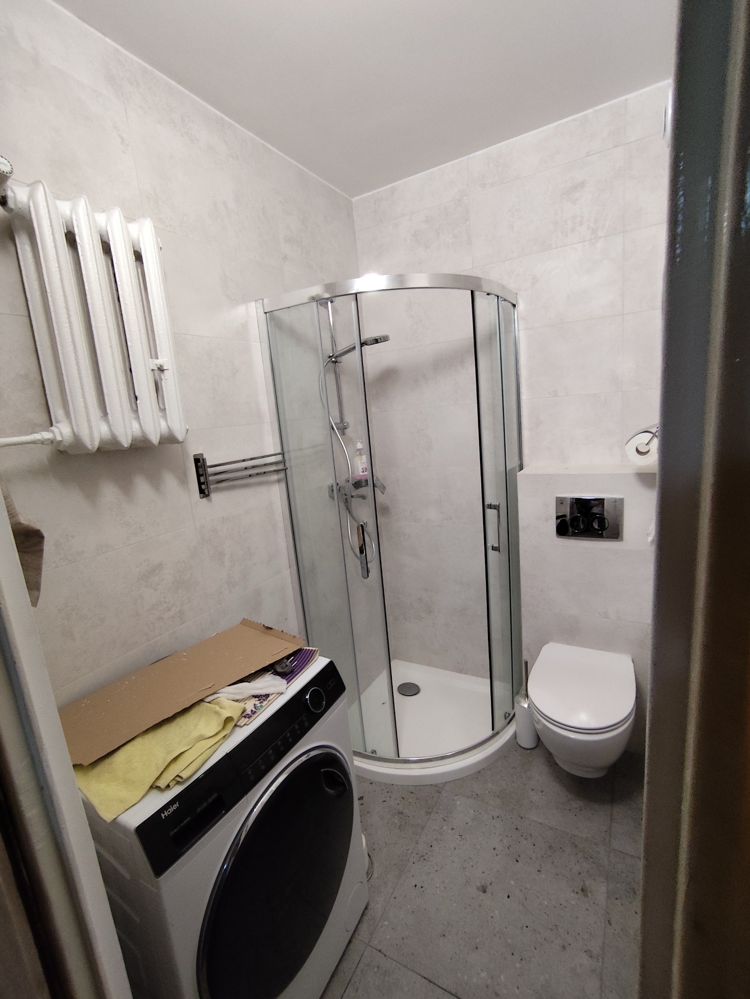 Wyremontowana łazienka z kabiną prysznicową, toaletą i pralką. Na ścianach jasne płytki imitujące kamień. Grzejnik żeberkowy. Ujęcie z perspektywy wejścia.