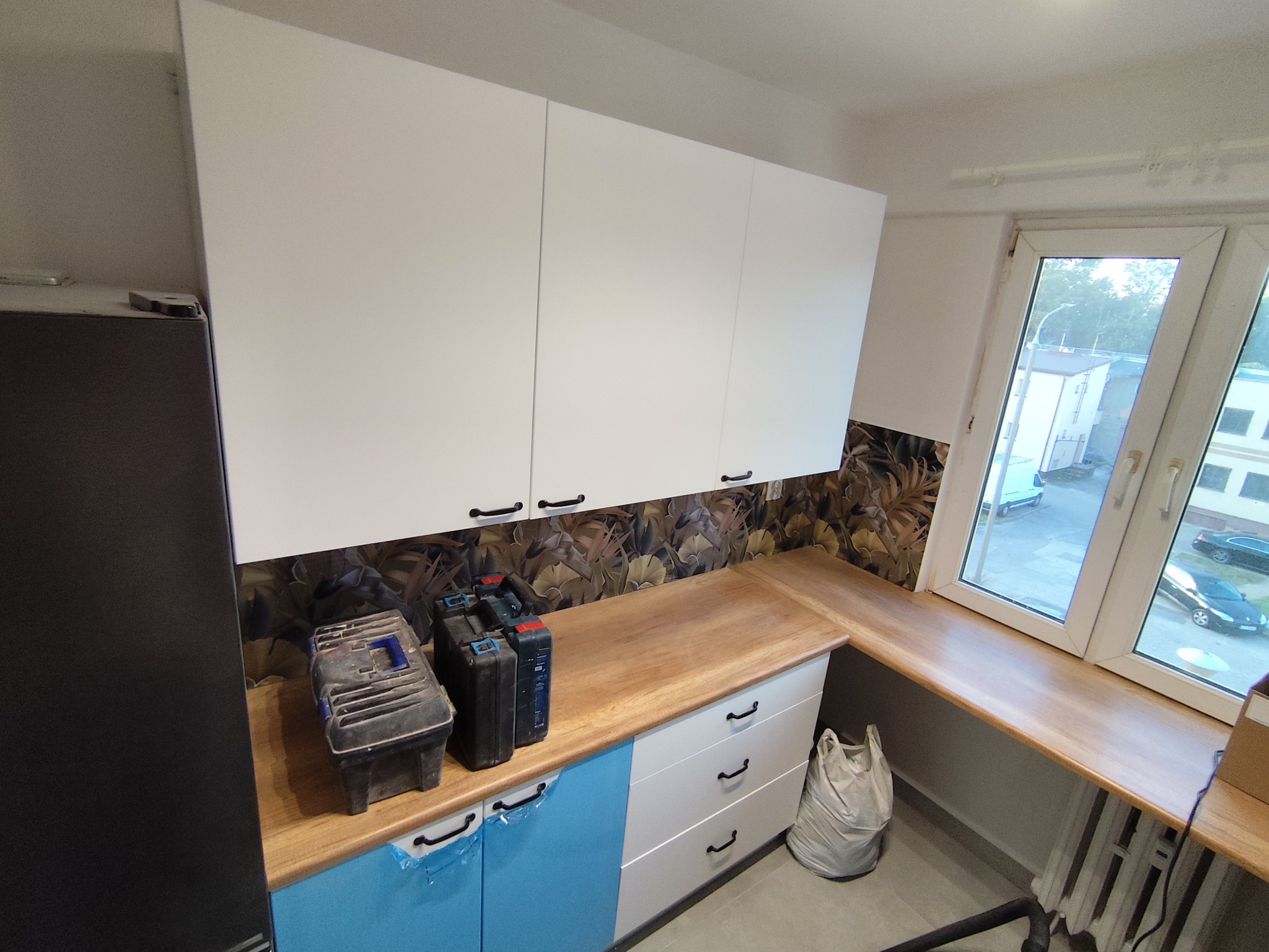 Nowoczesna kuchnia po remoncie: białe szafki wiszące z czarnymi uchwytami, drewniany blat, wzorzysta tapeta i okno z widokiem na zewnątrz. Widoczne narzędzia.