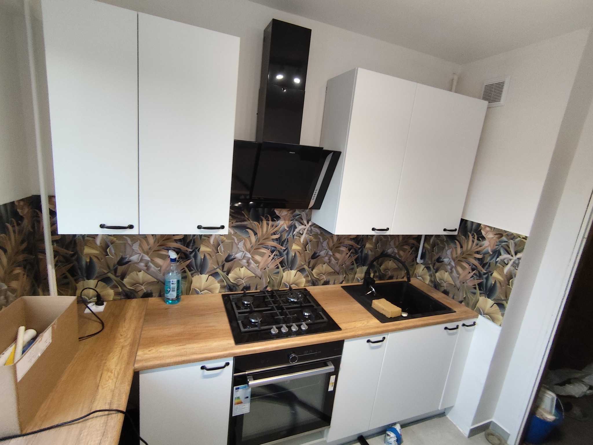 Nowoczesna kuchnia z białymi szafkami, drewnianym blatem i czarnym okapem. Na ścianie dekoracyjna tapeta w motywy roślinne. Widoczna płyta grzewcza i zlewozmywak.