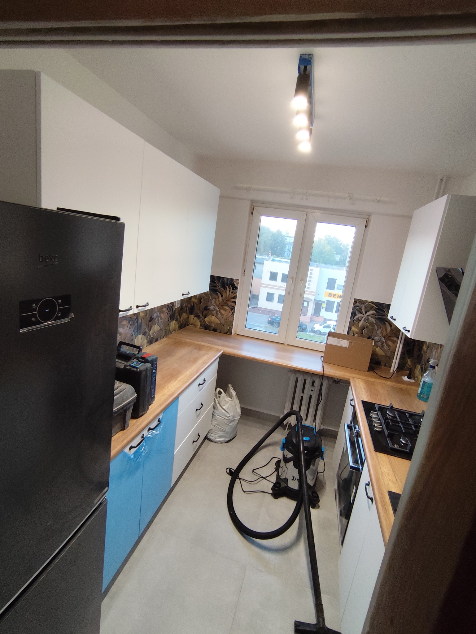 Kuchnia po remoncie: białe szafki z drewnianym blatem, niebieskie fronty dolne, wzorzysta ściana nad blatem. Widoczny odkurzacz i narzędzia, co sugeruje końcowy etap prac.