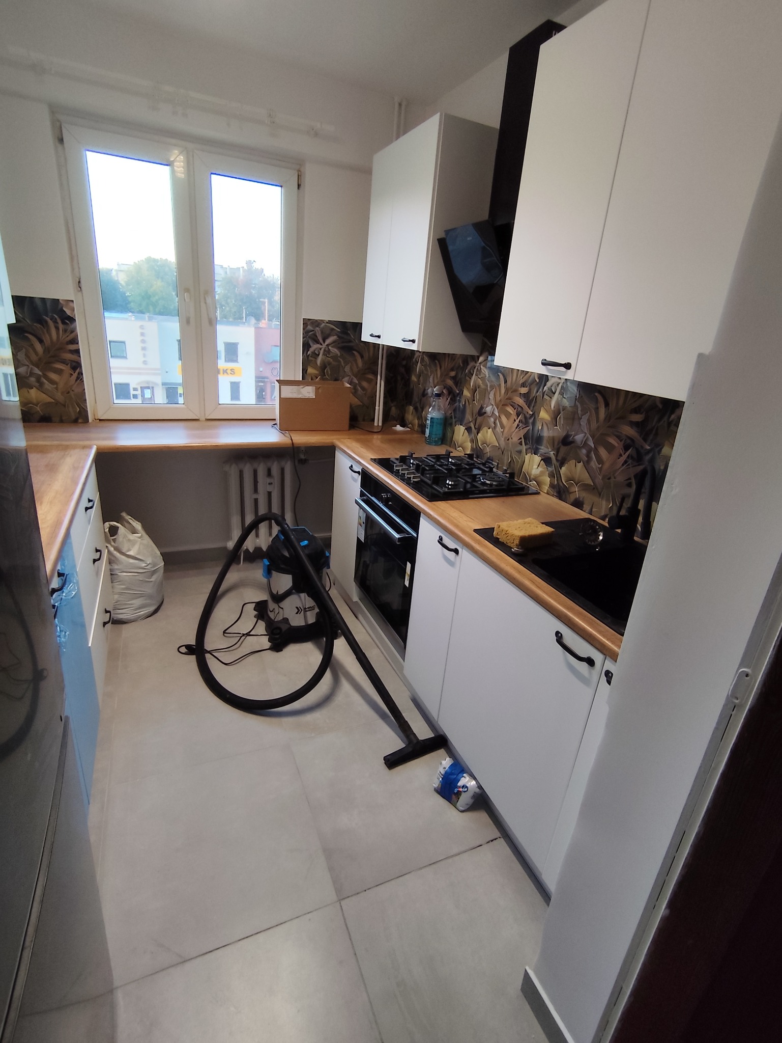 Kuchnia po remoncie: białe szafki z czarnymi uchwytami, drewniany blat, wzorzysta ściana nad blatem. Na podłodze odkurzacz i worek. Widok z okna na budynki.
