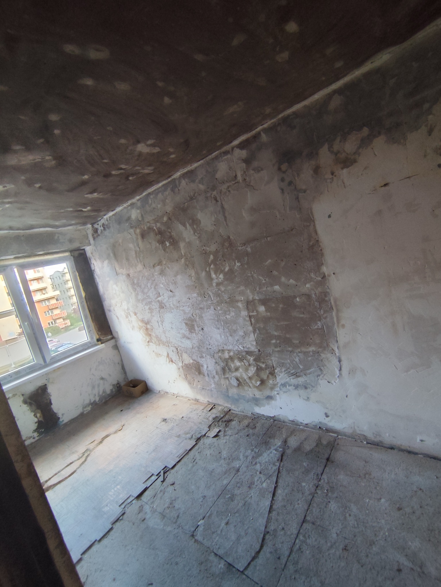 Pokój w trakcie prac przygotowawczych do remontu. Widoczne ślady po usuniętych tapetach, nierówności na ścianach i podłodze. Okno z widokiem na budynek mieszkalny.