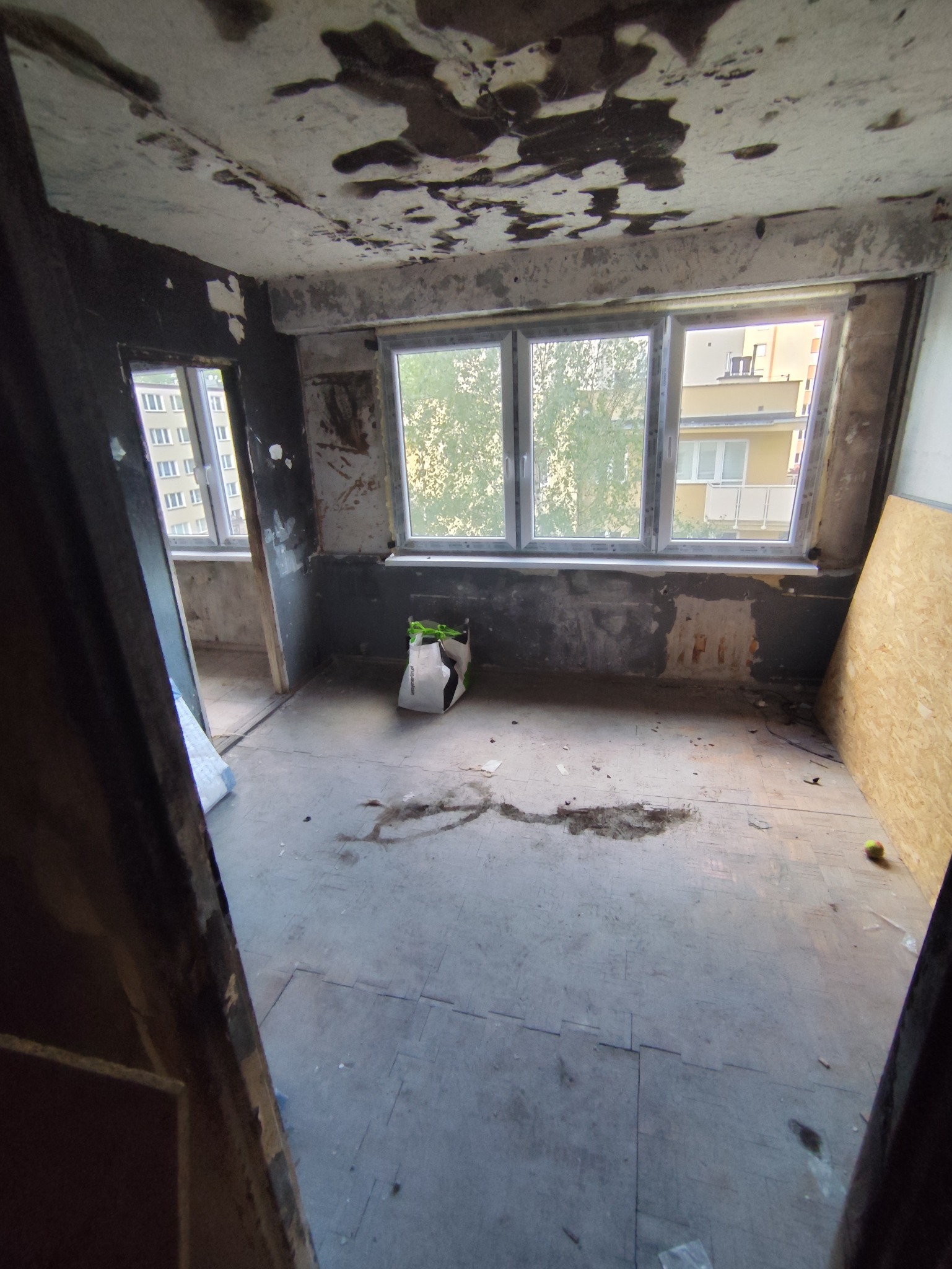 Wnętrze pokoju w trakcie remontu w Warszawie. Widoczne ślady po zalaniu na suficie, surowe ściany i podłoga. Torba z materiałami budowlanymi przy oknie.