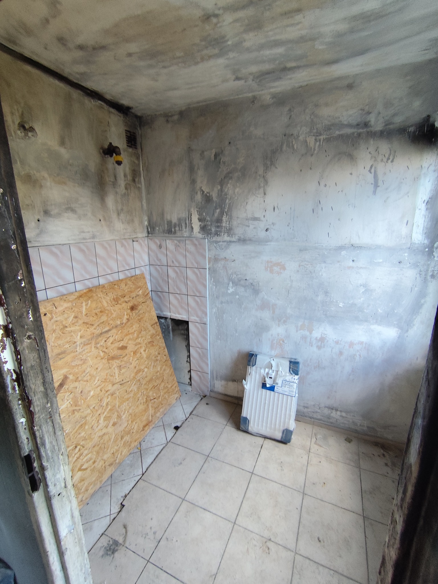 Wnętrze w trakcie remontu: surowe ściany, częściowo położone płytki, płyta OSB oparta o ścianę, nowy grzejnik w folii. Widoczne ślady po instalacjach.