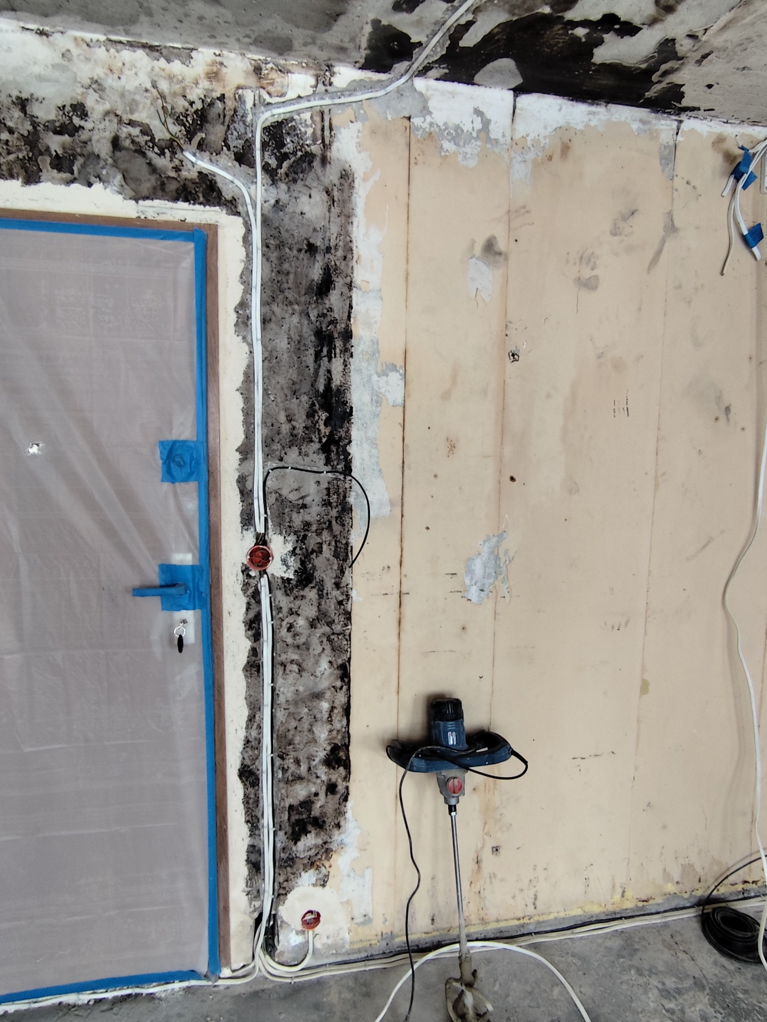 Przygotowanie do remontu: zabezpieczone drzwi folią, odsłonięta instalacja elektryczna, ślady wilgoci i grzyba na ścianie, mieszadło do zaprawy w pomieszczeniu.