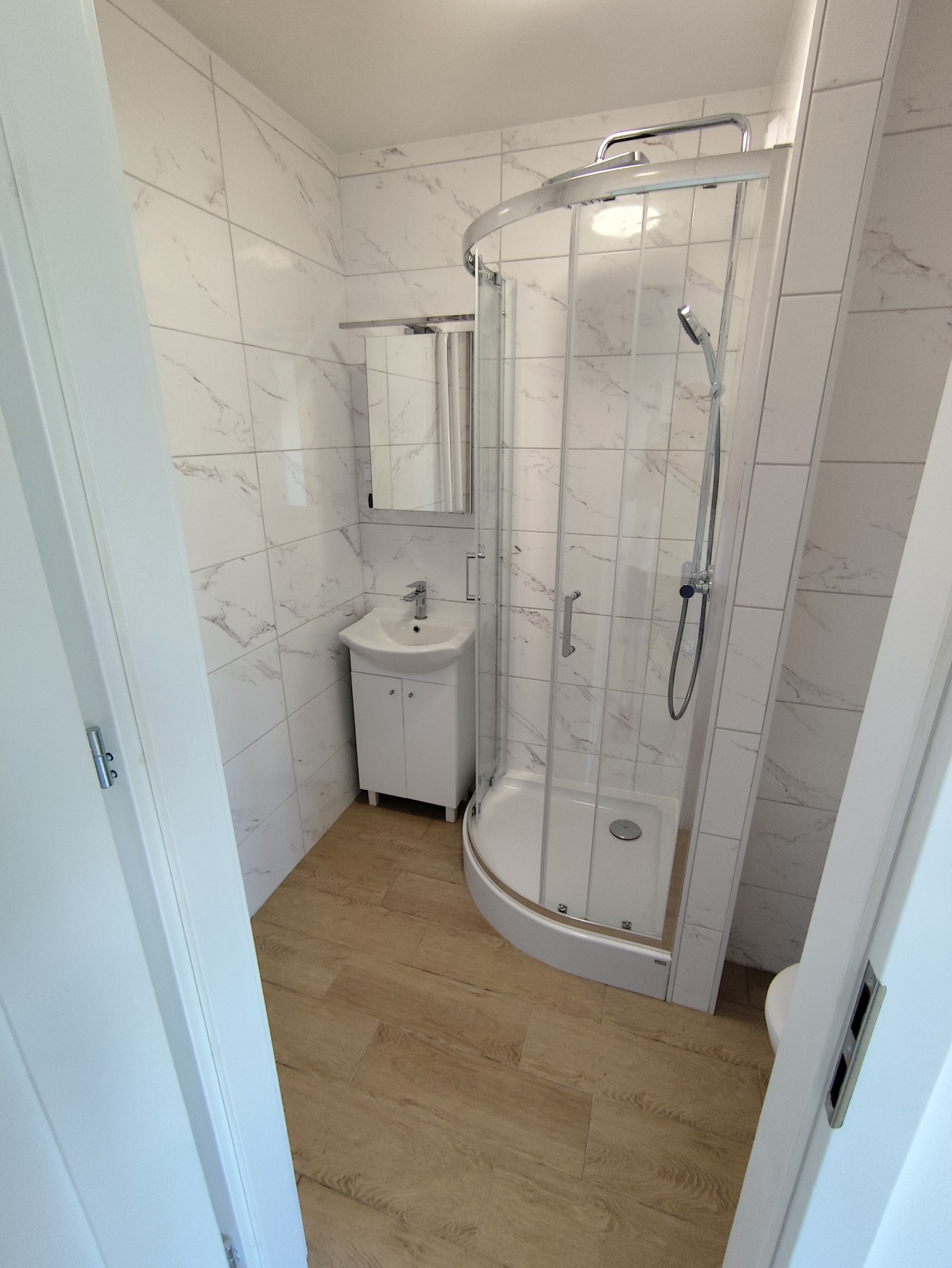 Jasna łazienka po remoncie: kabina prysznicowa z zaokrąglonymi drzwiami, umywalka z szafką, marmurowe płytki ścienne i podłoga imitująca drewno. Widok zza uchylonych drzwi.