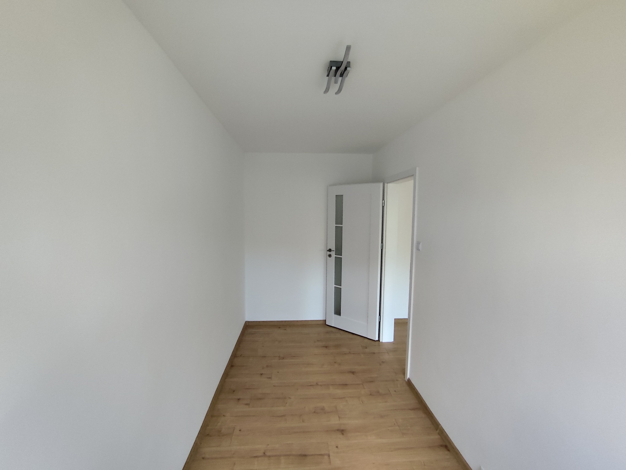 Jasne wnętrze z drewnianą podłogą i białymi ścianami. Widoczne drzwi z przeszkleniami i nowoczesna lampa sufitowa. Minimalistyczny design.