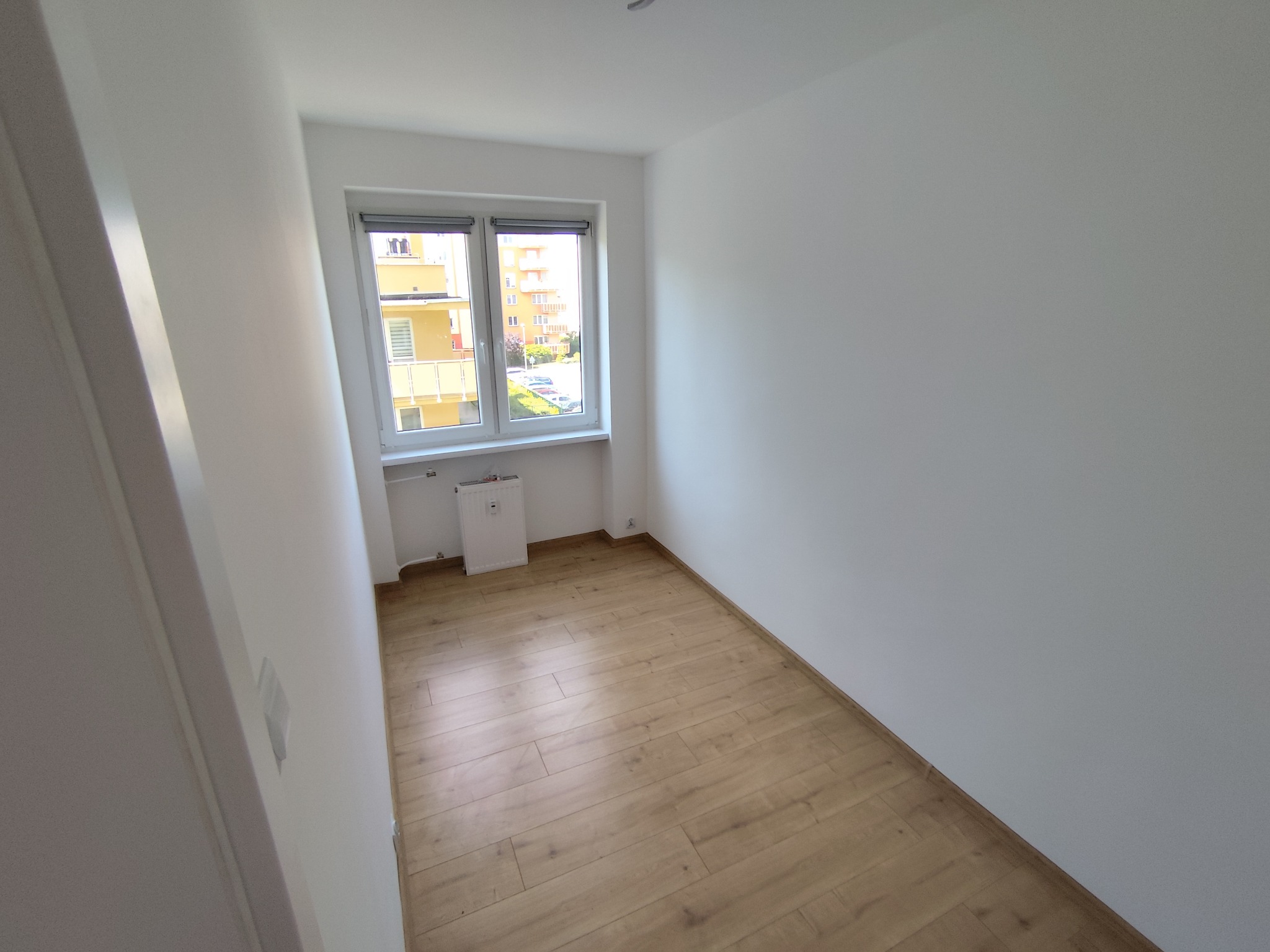 Jasne, puste pomieszczenie po remoncie z panelami podłogowymi i oknem z widokiem na budynek. Świeżo pomalowane ściany na biało. Grzejnik pod oknem.