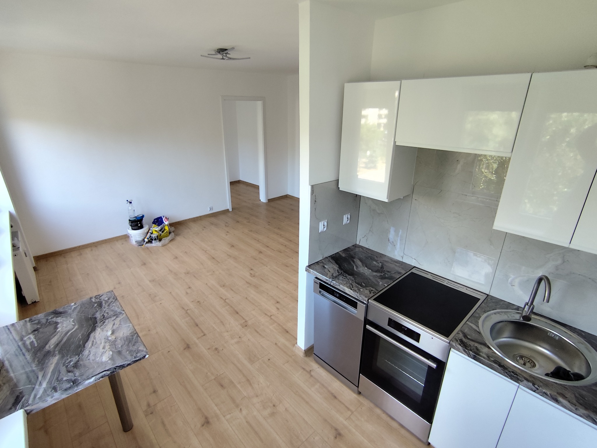 Jasne wnętrze po remoncie: białe ściany, panele podłogowe, nowoczesna kuchnia z marmurowym blatem i białymi szafkami. Widoczne ślady po malowaniu w pokoju.