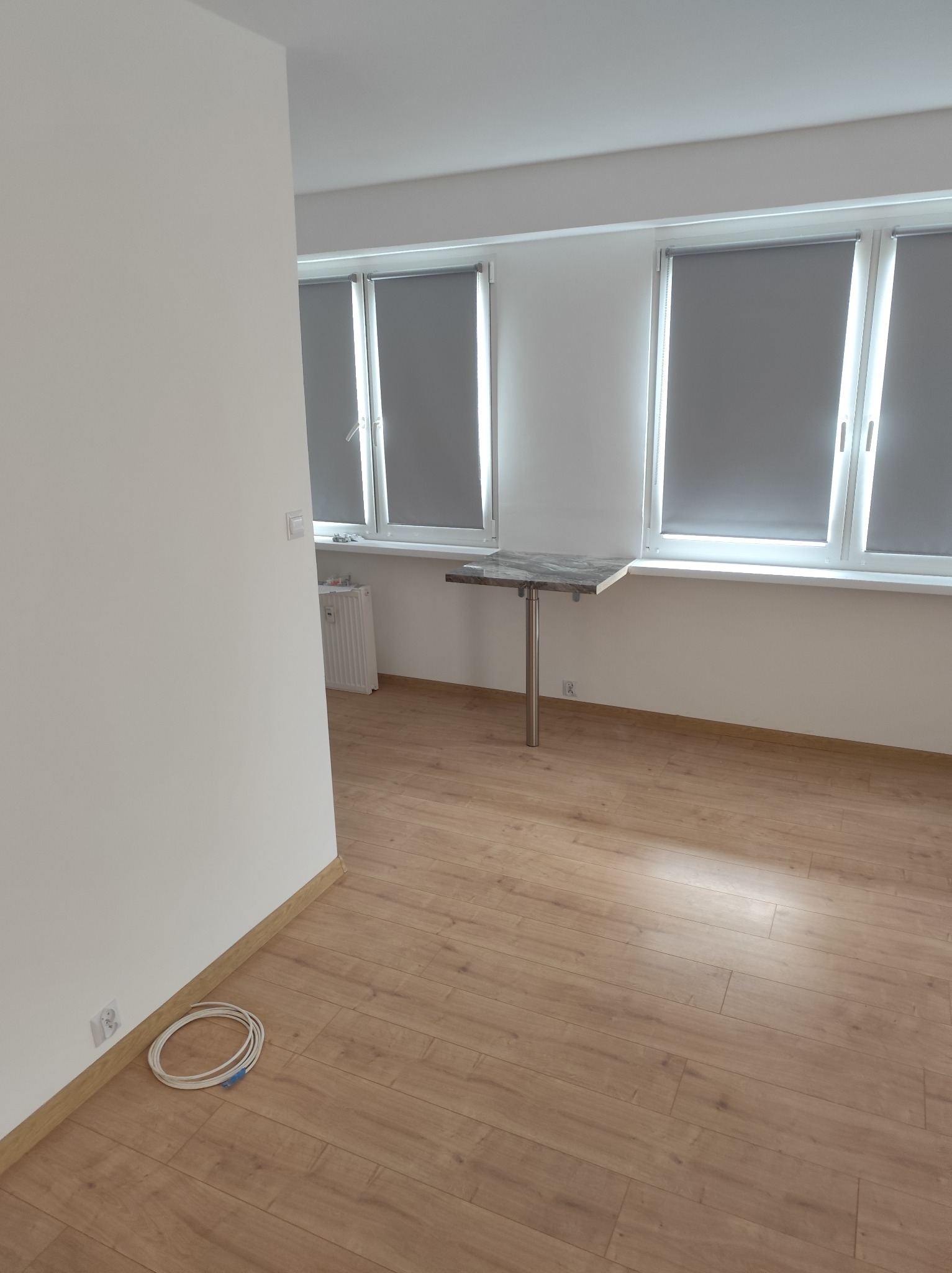 Jasne wnętrze po remoncie: parkiet, białe ściany, okna z roletami. Widoczne gniazdko i kabel przy podłodze, składany stolik przy oknie. Minimalistyczny styl.