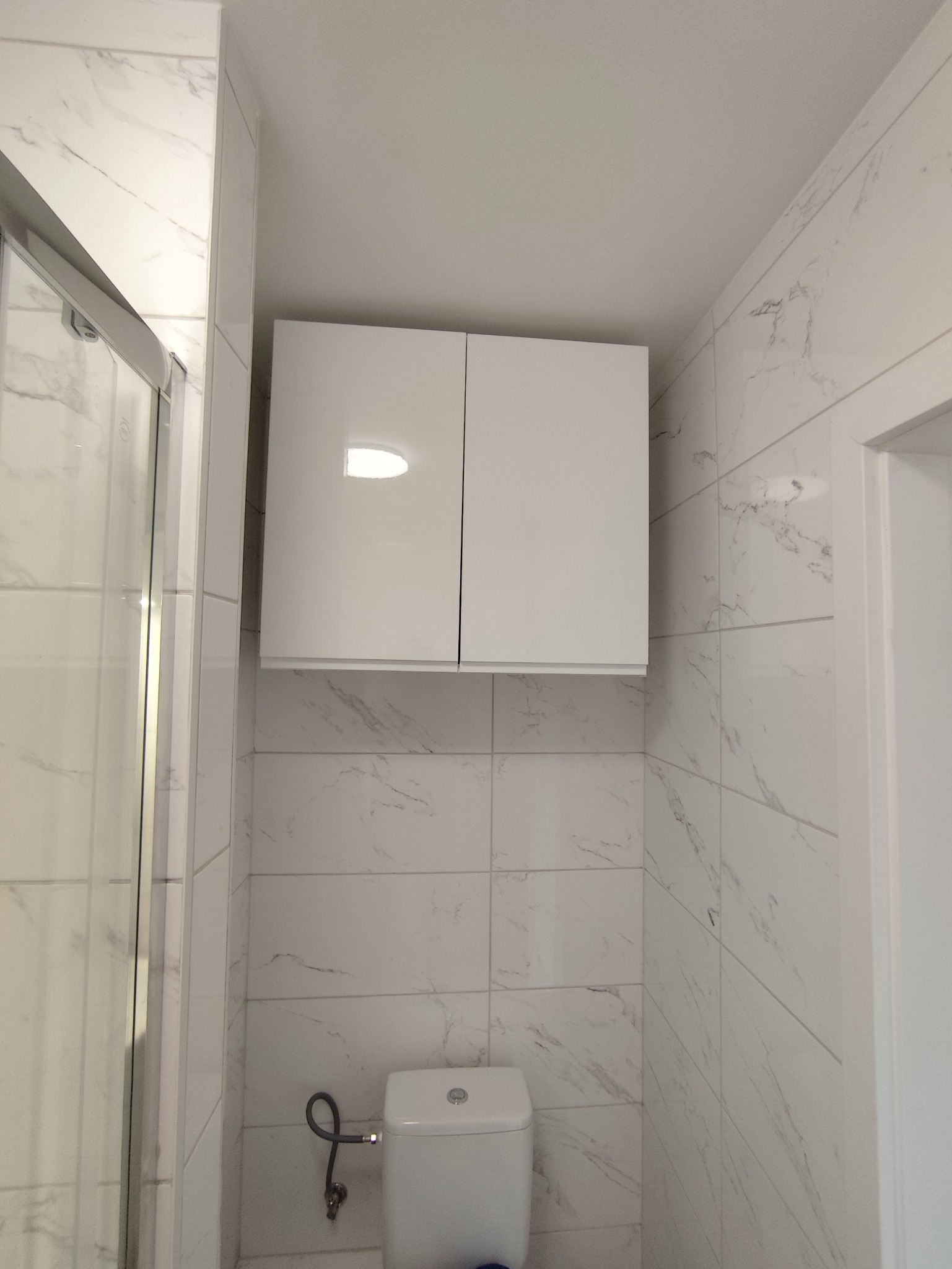 Biała, błyszcząca szafka nad toaletą w wykończonej łazience z marmurowymi płytkami. Widoczny fragment kabiny prysznicowej i drzwi. Minimalistyczny design.