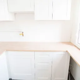 Nowa, jasna kuchnia z białymi szafkami i drewnianym blatem, widoczny zaw&oacute;r gazowy na ścianie i szary dywan na podłodze.