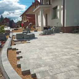 Trwa układanie szarej kostki brukowej na podjeździe przed domem; widoczne stosy kostki, pracownicy w trakcie pracy, zagęszczarka oraz fragment ułożonej krawędzi z podsypką żwirową.