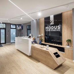 CustomMike - Nowoczesne biuro centrum rehabilitacji z recepcją o geometrycznym kształcie, drewnianymi panelami ściennymi i jasną podłogą, widoczne osoby pracujące.