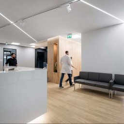 CustomMike - Nowoczesne wnętrze poczekalni biurowej z białą recepcją, drewnianymi panelami, szarą sofą i oświetleniem LED w suficie.