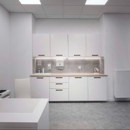 CustomMike - Jasne, minimalistyczne wnętrze gabinetu lekarskiego z białymi szafkami, drewnianym blatem, umywalką, radiatorem, biurkiem z laptopem i drukarką, oraz szarą podłogą.