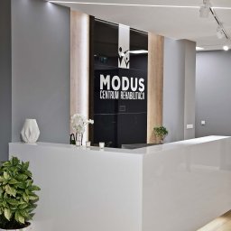 CustomMike - Nowoczesne biuro recepcyjne z białym, lakierowanym blatem, szarą ścianą i logo 'MODUS Centrum Rehabilitacji' na czarnym szkle, oświetlone liniowymi lampami LED i punktowymi reflektorami.
