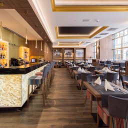 CustomMike - Eleganckie wnętrze restauracji z podświetlanym barem z onyksu, szarymi krzesłami barowymi i stolikami nakrytymi szarymi bieżnikami, widok na okna z widokiem na miasto.