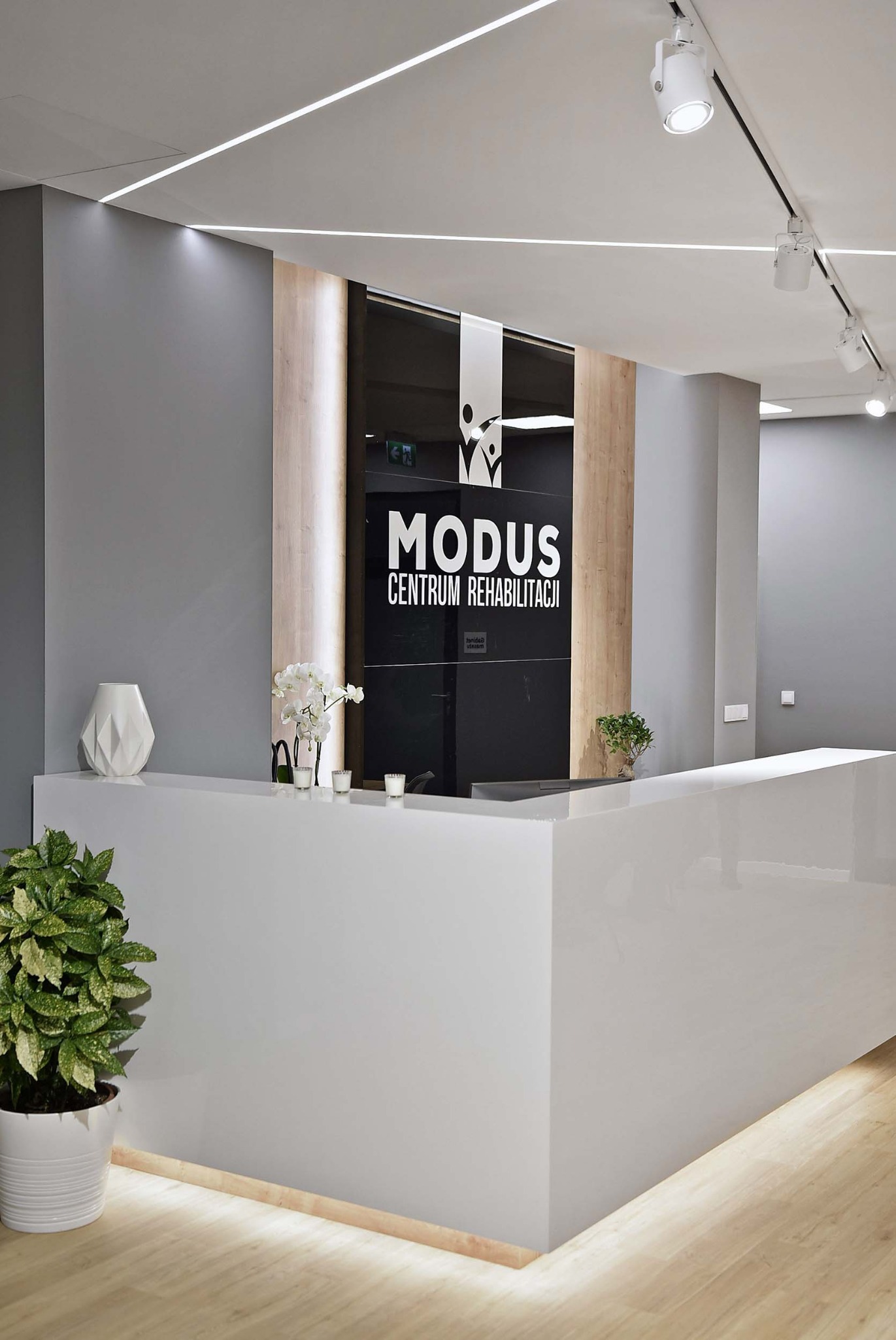 Nowoczesne biuro recepcyjne z białym, lakierowanym blatem, szarą ścianą i logo 'MODUS Centrum Rehabilitacji' na czarnym szkle, oświetlone liniowymi lampami LED i punktowymi reflektorami.