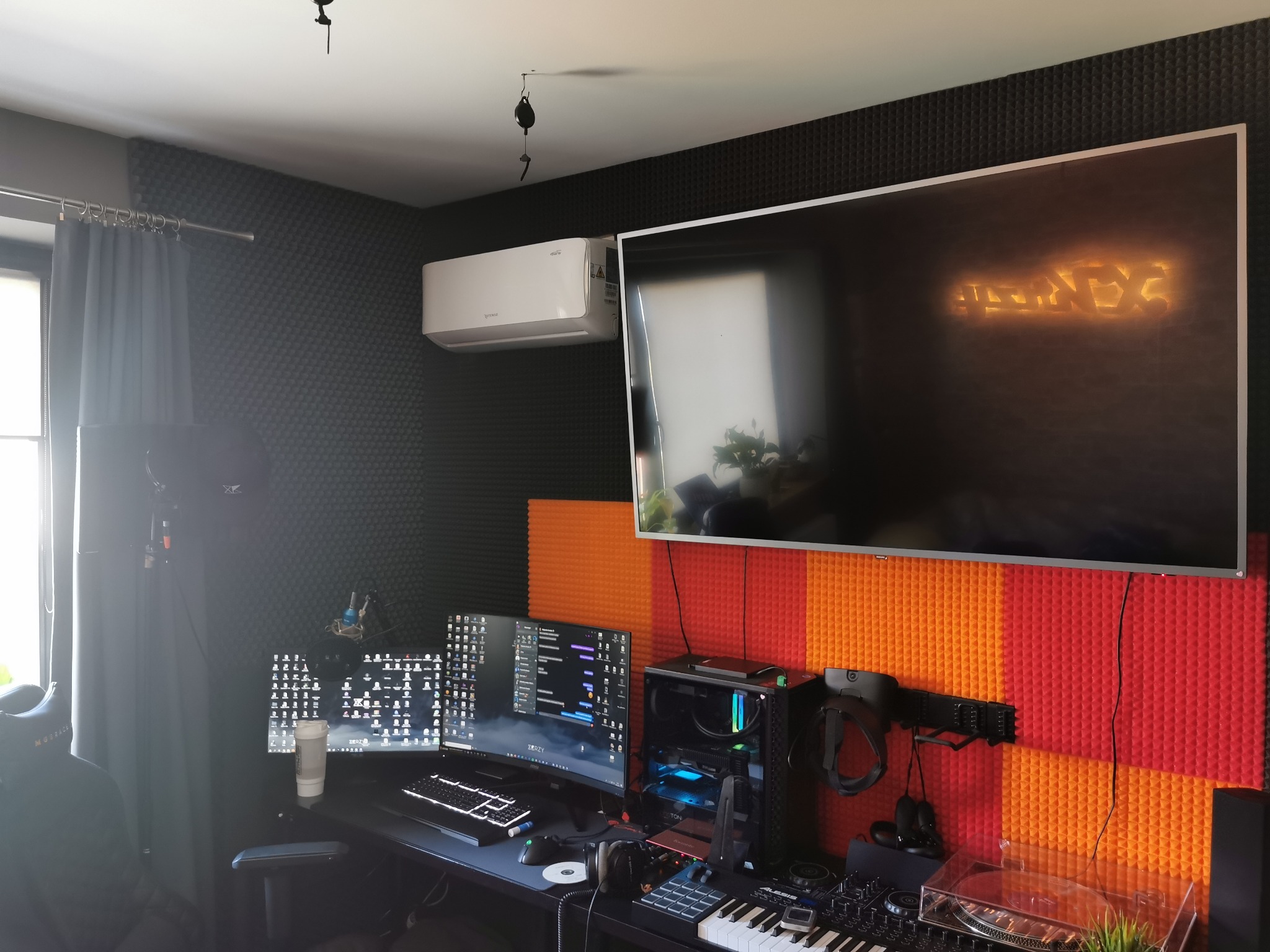 Wnętrze pokoju z zamontowaną klimatyzacją typu split, widoczną nad telewizorem, ściany wyłożone panelami akustycznymi w kolorze czarnym i czerwono-pomarańczowym, biurko z komputerem, monitorem...