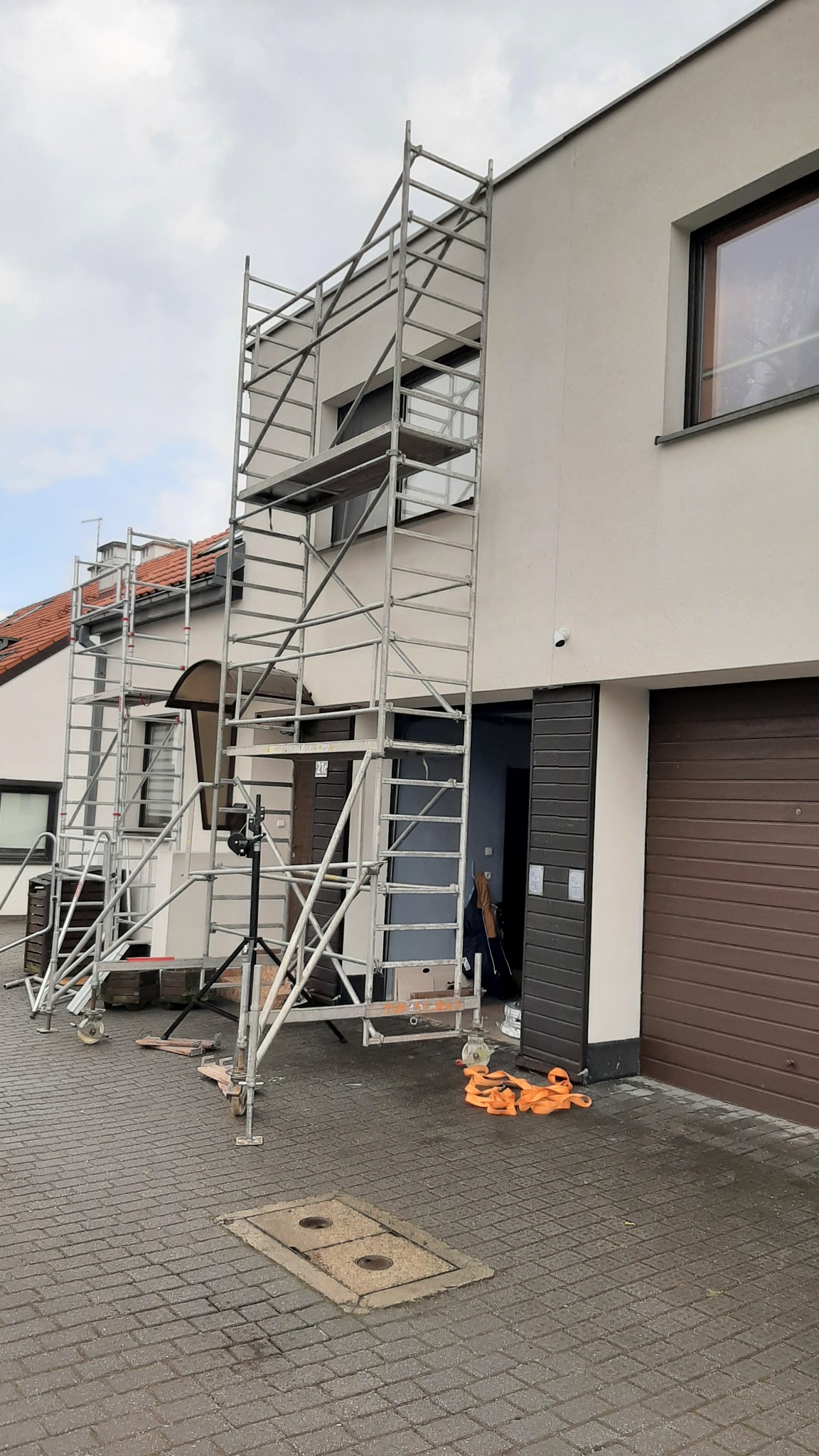 Dwa rusztowania aluminiowe ustawione przy ścianie dwukondygnacyjnego domu z garażem, widoczne pomarańczowe pasy zabezpieczające i sprzęt pomiarowy na statywie.