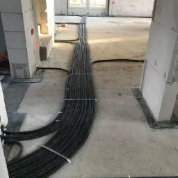 Wiązka czarnych, karbowanych rur instalacyjnych, prowadzonych po surowej posadzce wewnątrz budynku w stanie deweloperskim, zabezpieczonych metalowymi uchwytami.