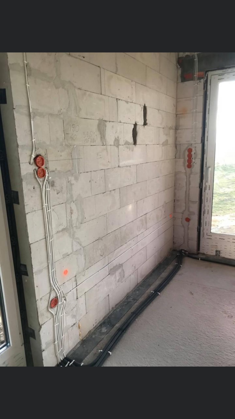 Surowy stan: Instalacja elektryczna w trakcie montażu w pomieszczeniu z białych bloczków, widoczne puszki instalacyjne, przewody w peszlach i czarne rury przy podłodze.