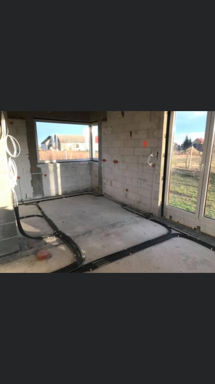 Surowy stan pomieszczenia z instalacją elektryczną: kable w peszlach ułożone wzdłuż ścian, puszki elektryczne zamontowane w ścianach z betonu komórkowego, widok przez okno na zabudowę jednorodzinną.