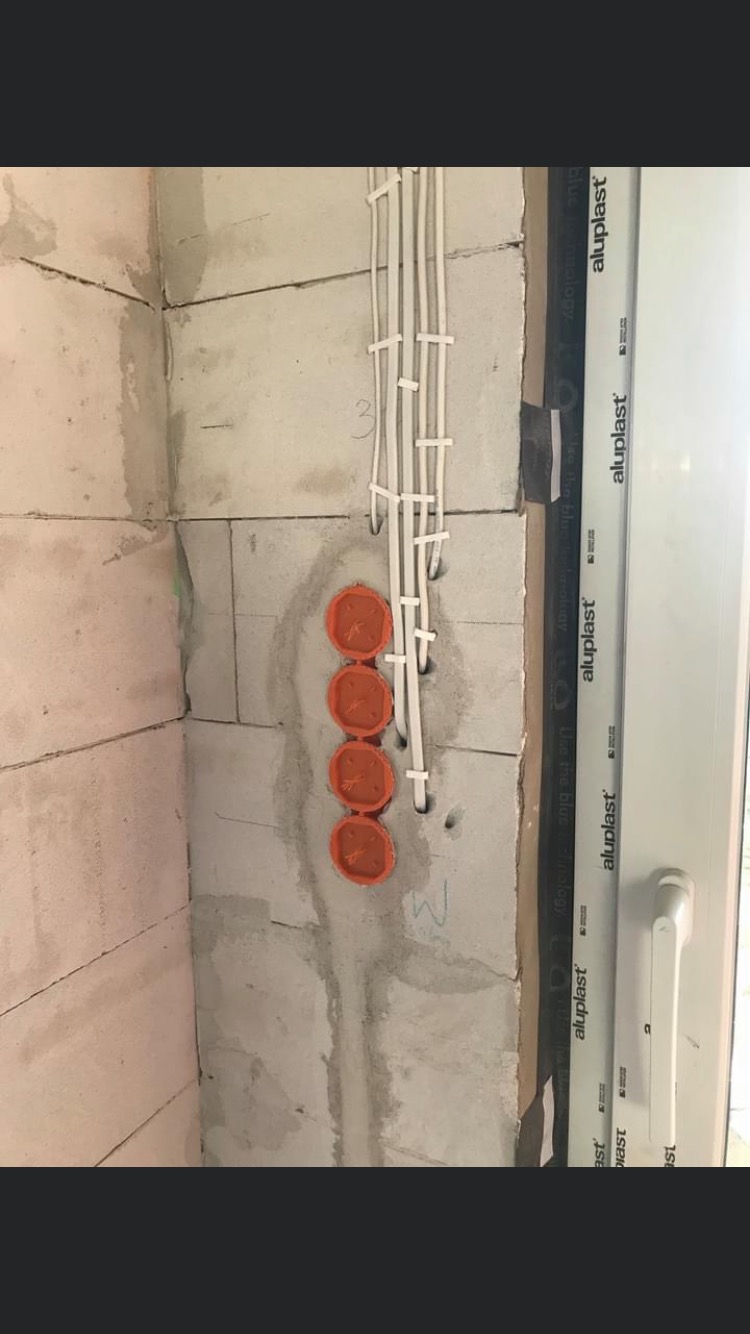 Ściana z betonu komórkowego z czterema pomarańczowymi puszkami pod gniazdka elektryczne i białymi kablami przymocowanymi uchwytami, widoczne ślady zaprawy.