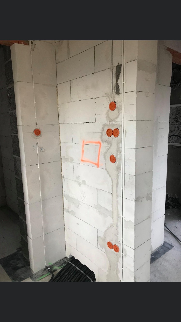 Instalacja elektryczna w surowym wnętrzu: widoczne puszki elektryczne w ścianach z betonu komórkowego, pionowe rury instalacyjne, oraz pomarańczowy kwadrat zaznaczony na ścianie.