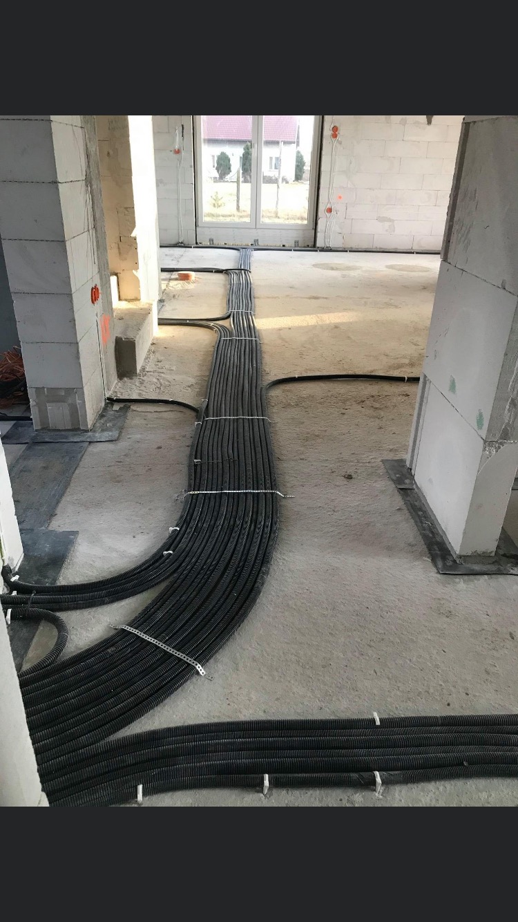 Wiązka czarnych, karbowanych rur instalacyjnych, prowadzonych po surowej posadzce wewnątrz budynku w stanie deweloperskim, zabezpieczonych metalowymi uchwytami.