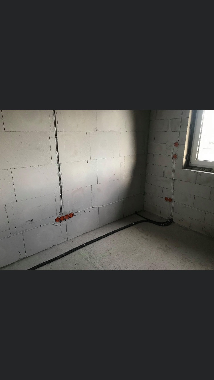 Surowy stan instalacji elektrycznej w pomieszczeniu: widoczne przewody i puszki podtynkowe na ścianach z bloczków betonowych, czarne rury instalacyjne na podłodze, okno w rogu.