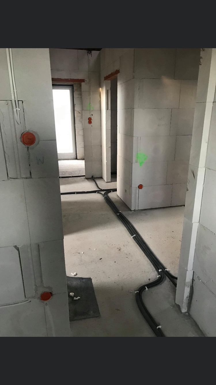 Wnętrze w stanie surowym z widocznymi kablami elektrycznymi ułożonymi wzdłuż ścian i podłogi, zabezpieczonymi białymi opaskami. Na ścianach widoczne pomarańczowe puszki instalacyjne i zielone...