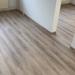 Jasna podłoga z paneli drewnopodobnych w pustym pokoju, widoczne grzejniki pod oknami i białe ściany, widok na sąsiednie pomieszczenie.