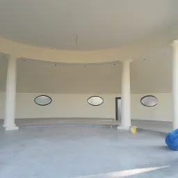 Okrągła, jasna sala z kolumnami w stylu klasycystycznym i owalnymi oknami; widoczne ślady prac wykończeniowych, wiadro farby i folia ochronna.